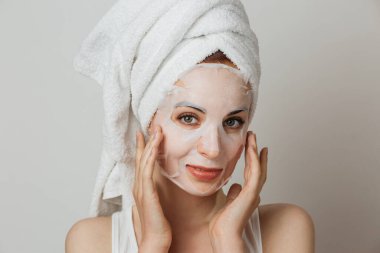 Stunning caucasian lady in towel applying paper mask sheet on face in studio. Young woman enjoying skin care procedures over grey background.
