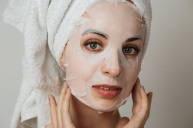 Attractive young woman with cotton mask on face looking at camera over grey background. Caucasian lady in towel and bare shoulders posing in studio.
