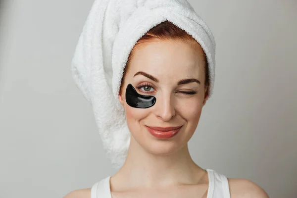 Young caucasian woman in towel with collagen black patch under eye over grey background. Beautiful female person using cosmetic for lifting and smoothing skin. Copy space. One eye closed.