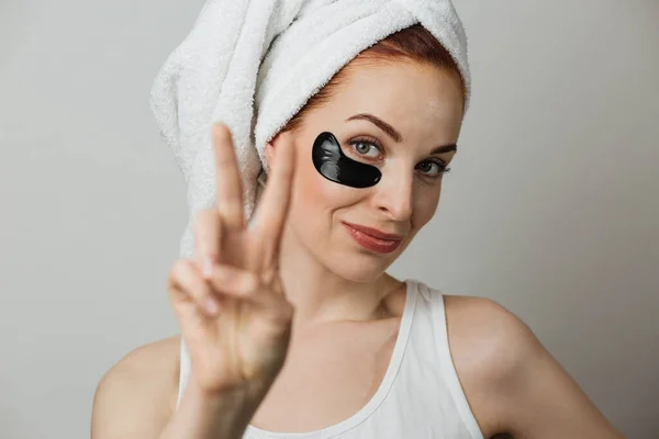 Close up face of adorable young woman in towel after shower, using black collagen patch for smoothing skin under eye showing sign of peace. Concept beauty treatments.