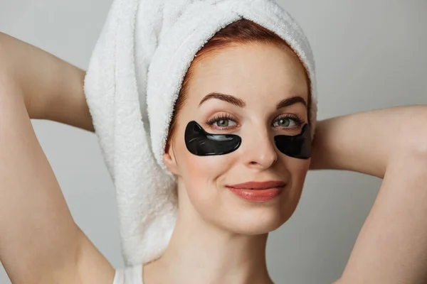 Charming young woman posing over grey background with black cosmetic patches under eyes. Adorable caucasian lady with bare shoulders enjoying her beauty anti aging procedures.