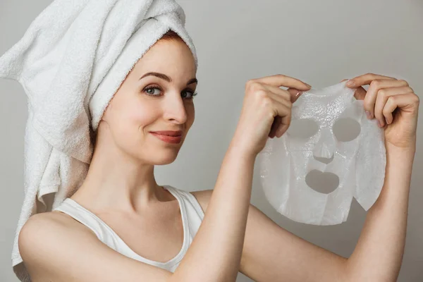 Attractive young lady with bare shoulders holding in hands cotton face mask over grey background. Portrait of happy woman using cosmetic for facial treatment.