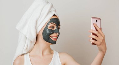 Attractive young woman with black cosmetic mask on her face holding smart phone. Portrait lady in towel over grey studio background.