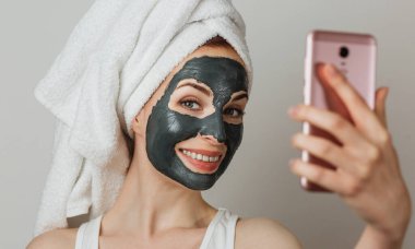 Smiling young woman with white clay mask on face holding using smart phone for selfie against grey background. Concept of aging process and cosmetic procedures.