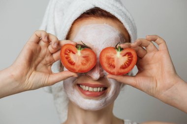 Portrait of caucasian young woman with white cosmetic mask on face and slices of tomato on eyes posing over grey background. Concept of skincare and regular procedure.
