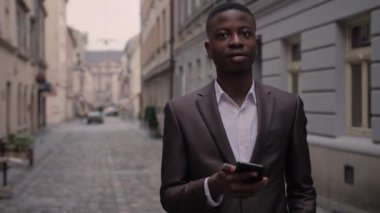 Şık takım elbiseli yakışıklı Afrikalı adam şehir caddesinde dururken modern akıllı telefondan e-postaları kontrol ediyor. Çalışma, başarı ve teknoloji kavramı.
