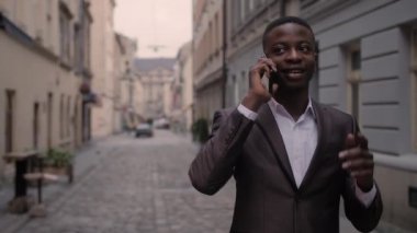İş kıyafetli Afrikalı adam şehir caddesinde yürürken mobil konuşma yapıyor. Yakışıklı genç adam gülümsüyor ve dışarıda konuşuyor..