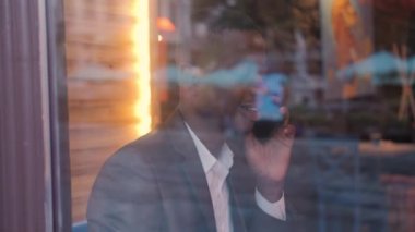 Serious afro american man in suit talking on mobile while sitting at cafe during a break. Young businessman having distance working conversation indoors.