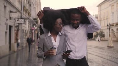 Şehir caddesinde yürürken ceketinin altında yağmurdan saklanan mutlu Afro-Amerikalı erkek ve kadın. İki iş ortağı iş toplantısına gidiyor..