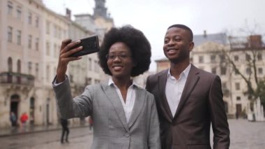 Başarılı afro-amerikan iş adamları şehir caddesinde yan yana durur ve akıllı telefondan selfie çekerler. Resmi giyinmiş genç adam ve kadın kamerada gülümsüyor ve el kol hareketi yapıyor..