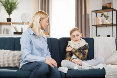 Pleasant 40-aged caucasian mother measuring temperature of her teen boy, which is sitting near her on soft couch with scarf around neck. Flu, covid-19 concept.