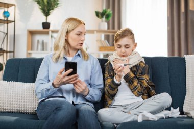Cold and flu, virus infection. Worried mother talking with doctor by phone, while her sick teen son with scarf around neck sitting on sofa at home with a thermometer after measuring the temperature.
