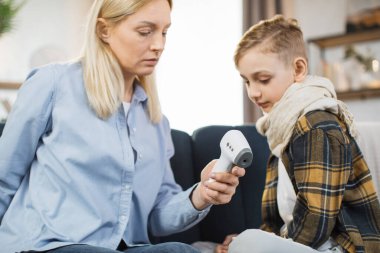 Mother taking care of her cute sick teen boy suffering from cold, checking temperature using infrared forehead thermometer gun, looking each other, sitting on the sofa.
