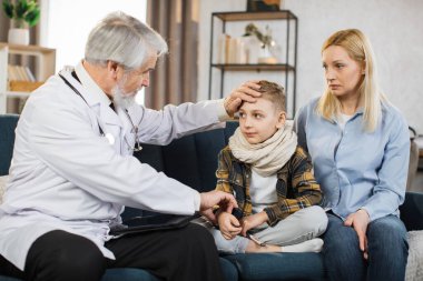 Front view of good-looking professional senior male caucasian doctor in medical coat, consulting his teen boy patient wihis mom, touching hand of child to check pulse, during home visit.