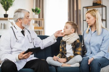 Mature doctor groping checking pain and swelling in the neck sore throat and cough thyroid nodule inflammation of the lymph nodes tonsillitis of sick male patient boy with his mom during home visit