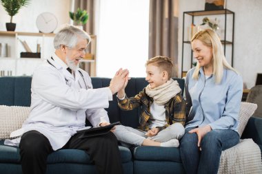 Mature man doctor giving high five during home visit of sick preschool boy patient sitting with mom, medical history or anamnesis, medical insurance contract.