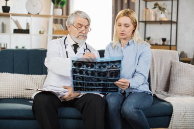 Medical care, home visit. Handsome mature confident male healthcare worker discussing the results of x-ray scan together with his young woman patient, worried about her health and condition.