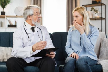 Front view shot of caring mature man doctor, consulting middle aged woman during homecare medical visit, making notes in clipboard. People healthcare concept during covid pandemic.