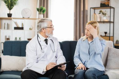 Grey hair bearded mature male doctor visits his pretty middle age patient at home. Therapist is taking notes, while young woman is complains of a terrible headache.