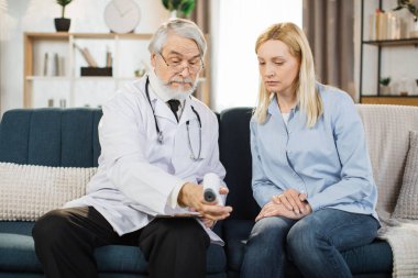 Caucasian male senior doctor using infrared thermometer gun, checking the body temperature of his sick woman patient, while visiting her at home.