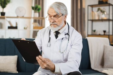 Confident grey haired doctor in eyewear and lab coat writing notes in flipchart while posing at hospital. Caucasian medical worker with stethoscope on neck standing indoors.