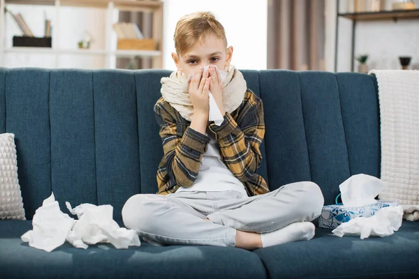 Coronavirus, pneumonia and flu concept. Front view of sick exhausted teen boy, sitting on the sofa at home, blowing running nose into paper handkerchief, having breathing difficulty, feeling bad.