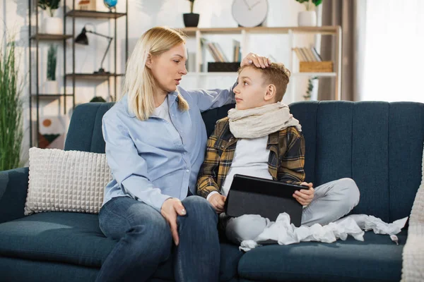 Family support during illness concept. Pleasant middle aged mother spending time at home together with her sick teen son, sitting on the sofa and using tablet pc.