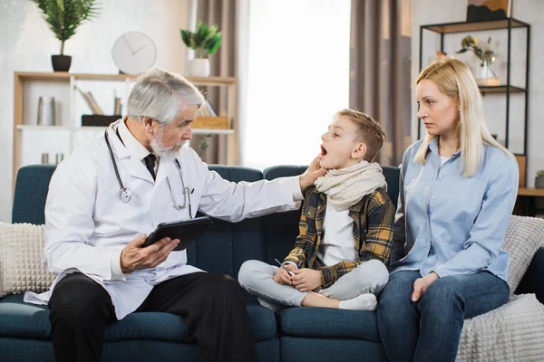 Experienced pleasant mature man doctor, examines throat of his sick preschool male patient, prescribing treatment during home visit. Doctor and patient with mother sitting on the couch at home.