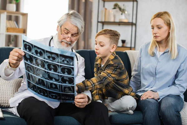 Pleasant mature man doctor supporting his preschool male patient with his mom during home visit, showing CT x-ray scan. Family doctor, little boy and mother , communicating and discussing treatment.