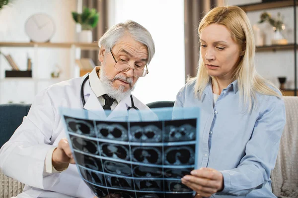 Senior bearded man confident doctor discussing the results of x-ray CT scan together with middle aged lady patient when visiting her at home for treatment control. Medicine concept, home visit.