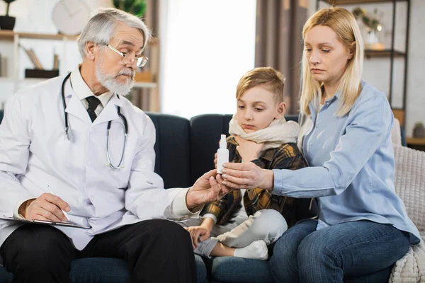 Experienced pleasant mature man doctor, giving nasal spray of his sick preschool male patient, prescribing treatment during home visit. Doctor and patient with mother sitting on the couch at home.