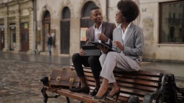 Resmi kıyafetli iki iş ortağı ahşap bankta oturup iş hakkında konuşuyorlar. Genç Afrikalı kadın panoya yazı yazıyor, yakışıklı adam dijital tablet kullanıyor..