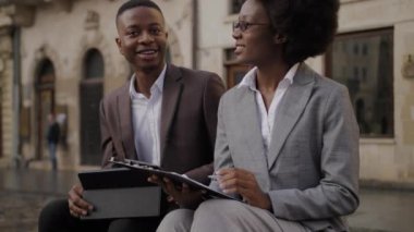 Elinde not panosu olan çekici bir kadın dijital tabletli genç bir adamın yanında ahşap bankta oturuyor. İki Afrikalı ortak dışarıda çalışma saatlerini tartışıyor..