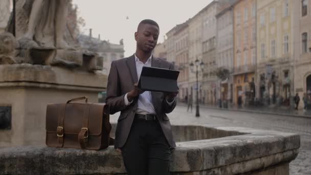身穿西装的黑人年轻人坐在城市街道的老喷泉上 一边用数字平板电脑 紧急工作在网上 现代技术概念 — 图库视频影像
