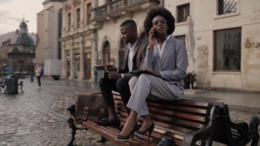 Erkek partner tahta bankta otururken ve dijital tablet kullanırken Afro-Amerikan kadın hareketli sohbetler yapıyor. Modern aletler ve uzaktan çalışma kavramı.