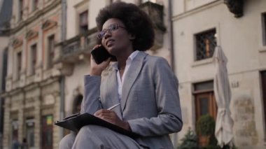 Şık giyinen, gözlüklü, akıllı telefondan konuşan ve panoya yazı yazan başarılı bir iş kadını. Afro-Amerikalı kadın bankta oturuyor ve dışarıda çalışıyor..