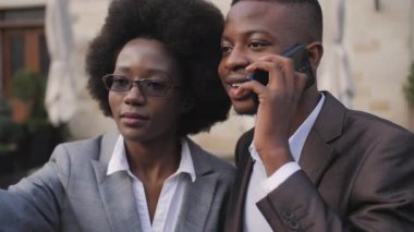 Gülümseyen Afrikalı bir adam ve takım elbiseli bir kadın şehrin caddesinde birlikte durup modern akıllı telefondan selfie çekiyorlar. Dışarıda mutlu ortaklar.