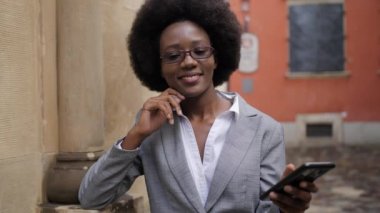 Attractive business african woman standing on city street with clipboard and modern smartphone in hands. Busy lady in suit and eyeglasses solving working issues online.