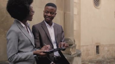 Handsome african businessman pointing with finger on clipboard that holding attractive woman. Two partners analyzing some documents on street.