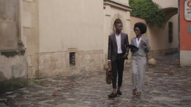 Resmi giyinmiş iki Afrikalı iş adamının uzun bir portresi. Şehir caddesinde yürüyor ve konuşuyor. Kahverengi evrak çantası taşıyan yakışıklı genç adam, elinde pano tutan kadın..