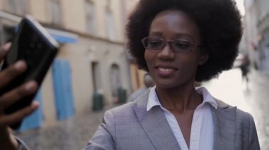 Çekici Afrikalı iş kadını modern akıllı telefondaki video görüşmesi sırasında gülümsüyor ve el kol hareketi yapıyor. Gözlüklü ve takım elbiseli mutlu kadın şehir caddesinde duruyor ve cep telefonuyla konuşuyor..