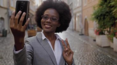 Çekici Afrikalı iş kadını modern akıllı telefondaki video görüşmesi sırasında gülümsüyor ve el kol hareketi yapıyor. Gözlüklü ve takım elbiseli mutlu kadın şehir caddesinde duruyor ve cep telefonuyla konuşuyor..