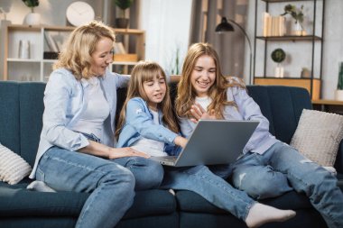 Happy family of mother and two cute daughters sitting together on comfortable couch, using laptop, resting and spend free time at home, recording video in living room.