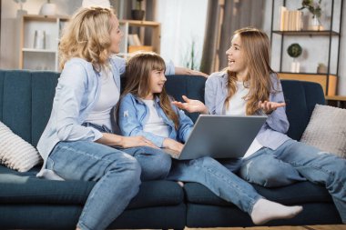 Happy family with two children having good time using laptop. Young smiling mother with cute blond daughters sitting on sofa at home looking at computer screen and watching video.