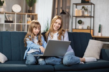 Happy sisters or friends two cute blond girls sitting together on comfortable couch, using laptop, holding video call with father or greeting grandparents, recording video in living room.