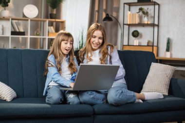 Happy sisters or friends two cute blond girls sitting together on comfortable couch, using laptop, holding video call with father or greeting grandparents, recording video in living room.