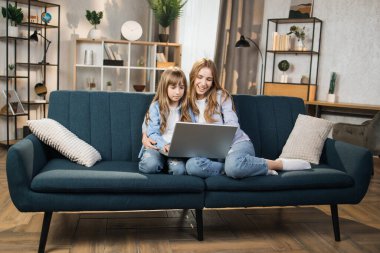 Caucasian cute female children sitting on couch using laptop. Two sisters enjoying distant talk by video call, spend time in internet buying, e-commerce users, watching cartoons or video film.
