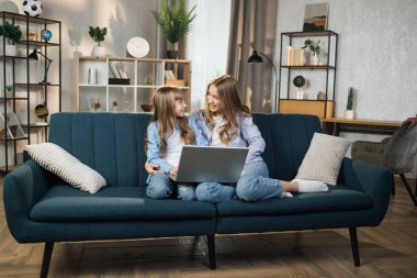 Caucasian cute female children sitting on couch using laptop. Two sisters enjoying distant talk by video call, spend time in internet buying, e-commerce users, watching cartoons or video film.
