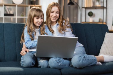 Caucasian cute female children sitting on couch using laptop. Two sisters enjoying distant talk by video call, spend time in internet buying, e-commerce users, watching cartoons or video film.