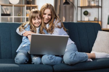 Caucasian cute female children sitting on couch using laptop. Two sisters enjoying distant talk by video call, spend time in internet buying, e-commerce users, watching cartoons or video film.